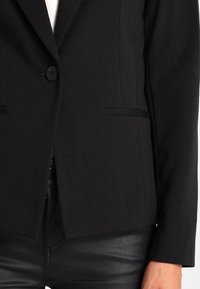 Černý upravený blazer s jedním knoflíkem, klopovými límci a dvěma předními kapsami. Hladká tkanina a strukturovaný střih.