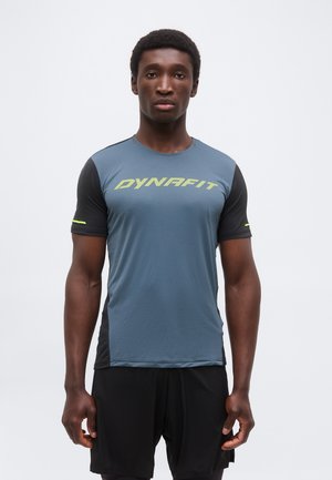 Férfi áll, előre néz, kék-fekete Dynafit rövid ujjú sportfelsőt és fekete rövidnadrágot visel, egyszínű háttér előtt.