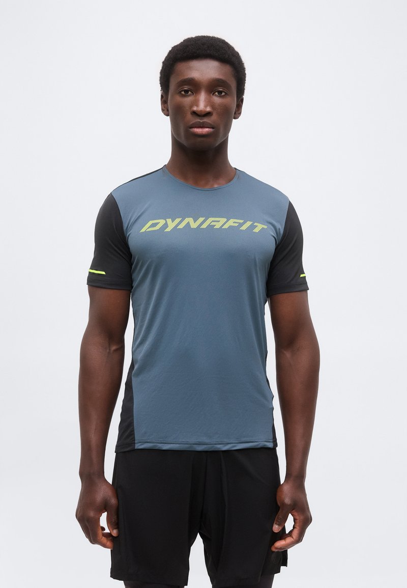 Άνδρας που στέκεται βλέποντας μπροστά, φοράει μπλε και μαύρο αθλητικό κοντομάνικο μπλουζάκι Dynafit και μαύρα σορτς σε απλό φόντο.