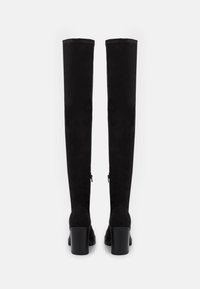 Bottes hautes en daim noir jusqu'à la cuisse avec un design droit, talons carrés épais, fermetures éclair latérales, présentant une texture lisse et un style minimaliste.