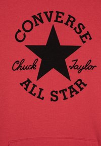 Crvena dukserica sa velikom crnom zvijezdom i natpisom "Converse Chuck Taylor All Star" u podebljanom, kružnom rasporedu oko nje.
