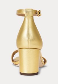 Zlaté sandály na vysokém podpatku s hladkým metalickým povrchem, pásem kolem kotníku a hranatým blokovým podpatkem. Hladká textura, minimalistický design.