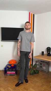 Maglietta grigia a maniche corte con design grafico, abbinata a jeans scuri e scarpe scure. Ambientazione interna con una pianta e accenti di colore sulla parete.