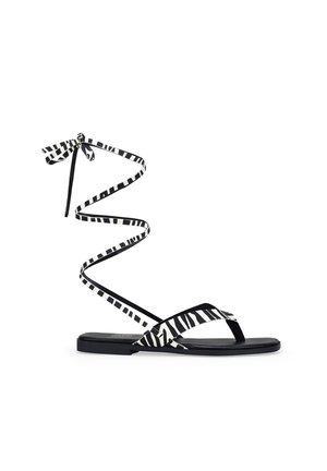 Sandalias planas negras que presentan una tira de tela a rayas en blanco y negro que se enrolla alrededor del tobillo y un diseño minimalista.