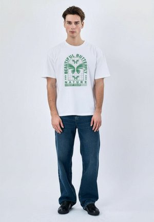 Junger Mann steht, trägt weißes T-Shirt mit grünem Schmetterlingsmotiv und blaue Jeans, schwarze Schuhe, einfarbiger heller Hintergrund.