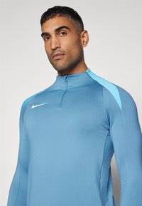 Világoskék sportfelső félig zipzárral, hosszú ujjakkal és kontrasztos sötétkék vállbetétekkel. A mellkason a Nike logó látható.