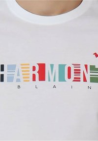 T-shirt bianco con testo multicolore "HARMONIE" e "BLAIN" in diversi stili di carattere, accompagnato da una piccola icona rossa. Tessuto di cotone liscio.