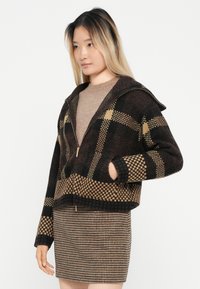 Braun und schwarz kariertes Zip-Up-Jackett mit Kapuze. Verfügt über kontrastierende Karomuster und einen verkürzten Schnitt. Getragen über einem hellbraunen Oberteil.