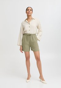 Helle beigen Hemd mit Knöpfen und langen Ärmeln, kombiniert mit olivgrünen Shorts, die einen elastischen Bund und seitliche Taschen haben. Die Heels runden das Outfit ab.