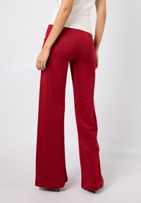 Røde, vide bukser med brede ben laget av et glitrende stoff, med høyt liv og glatt tekstur. Bakfra vises en lett utsving ved kanten.