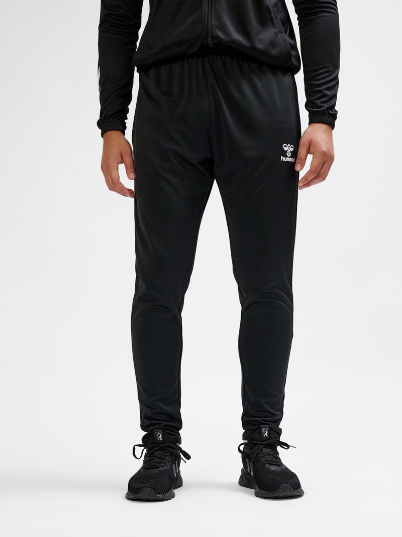 Uomo che indossa una giacca sportiva nera, pantaloni da jogging aderenti neri con logo sulla coscia e scarpe sportive nere, in piedi su sfondo bianco.