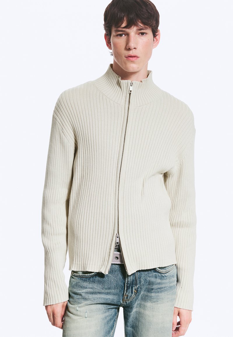 Jeune homme portant un pull zippé côtelé couleur crème et un jean bleu délavé, debout devant un fond blanc uni.