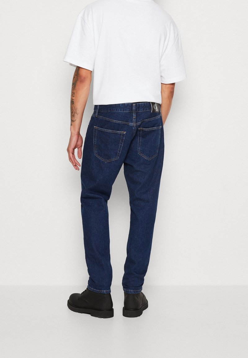 Vaqueros de denim azul oscuro con corte recto, cinco bolsillos y costuras en contraste; combinados con una camiseta blanca simple y botas negras.