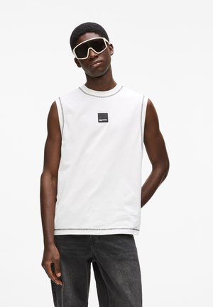 Mlad moški, ki nosi preveliko belo majico brez rokavov, črne kavbojke in velike bele sončna očala s črnimi okvirji, stoji z eno roko za hrbtom.