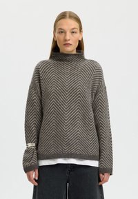 Maglione a zigzag marrone e crema con collo alto, maniche larghe, maglia strutturata e indossato sopra una camicia bianca.