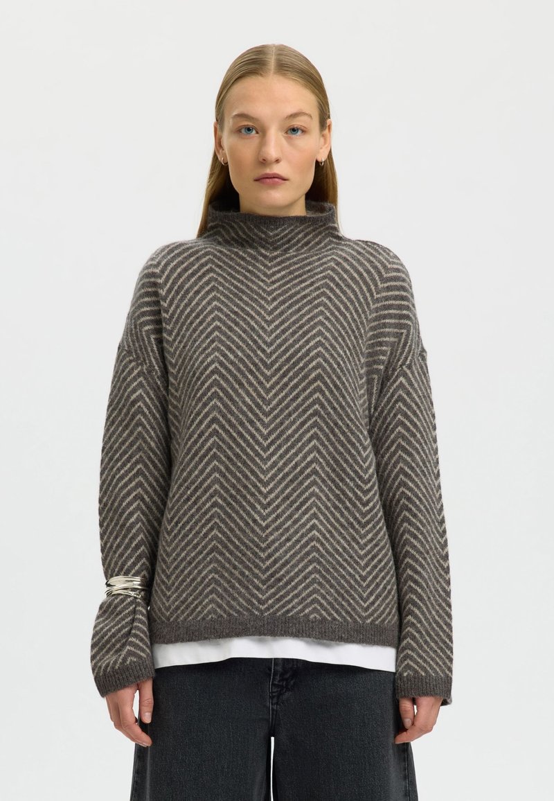 Maglione a zigzag marrone e crema con collo alto, maniche larghe, maglia strutturata e indossato sopra una camicia bianca.