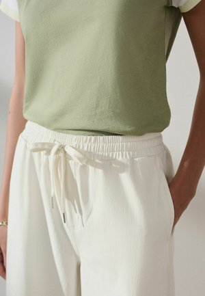 Donna con indosso una canotta verde chiaro infilata in pantaloni bianchi con coulisse, con le mani in tasca, vista dalle spalle ai fianchi.