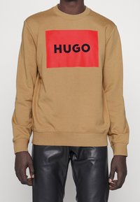 Hombre con sudadera color camello con un gran cuadrado rojo y texto negro "HUGO", combinado con pantalones de cuero negros.