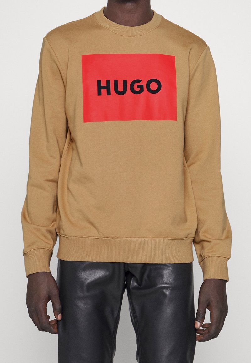 Hombre con sudadera color camello con un gran cuadrado rojo y texto negro "HUGO", combinado con pantalones de cuero negros.