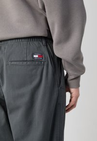 Szürke nadrág sima anyagból, elasztikus derékkal, oldalsó zsebekkel, és Tommy Jeans logóval a hátsó részen.