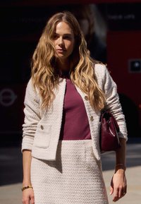 Texturierte cremefarbene Blazerjacke mit goldenen Knöpfen über einem taillierten burgunderfarbenen Oberteil, kombiniert mit einem cremefarbenen Rock und einer kleinen burgunderfarbenen Handtasche.