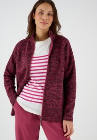 Chaqueta cremosa con cremallera en color marrón burdeos y tejido texturizado, con cuello alto y bolsillos. Debajo, una camisa blanca con rayas rosas.
