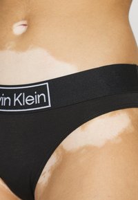 Fekete pamutzokni vastag derékpánttal, amelyen a "Calvin Klein" logó látható. A bőr a derékpánt közelében eltérő pigmentációt mutat.