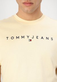 Gelbes Baumwoll-T-Shirt mit schwarzem "TOMMYJEANS"-Logo und einem kleinen roten und blauen Flaggenakzent darunter. Klassisches Rundhalsausschnitt-Design.