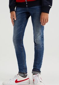 Smal blå denimjeans med ljus blekning och whiskering, med en uppvikt kant vid ankeln, i kombination med vita sneakers och röda accenter.