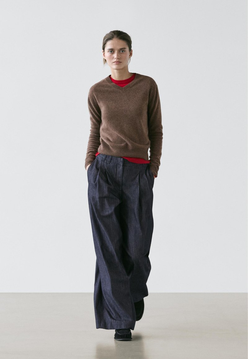 Pull en laine marron à col en V superposé sur une chemise rouge, associé à un pantalon ample bleu foncé plissé. Design simple et moderne, tissu texturé.