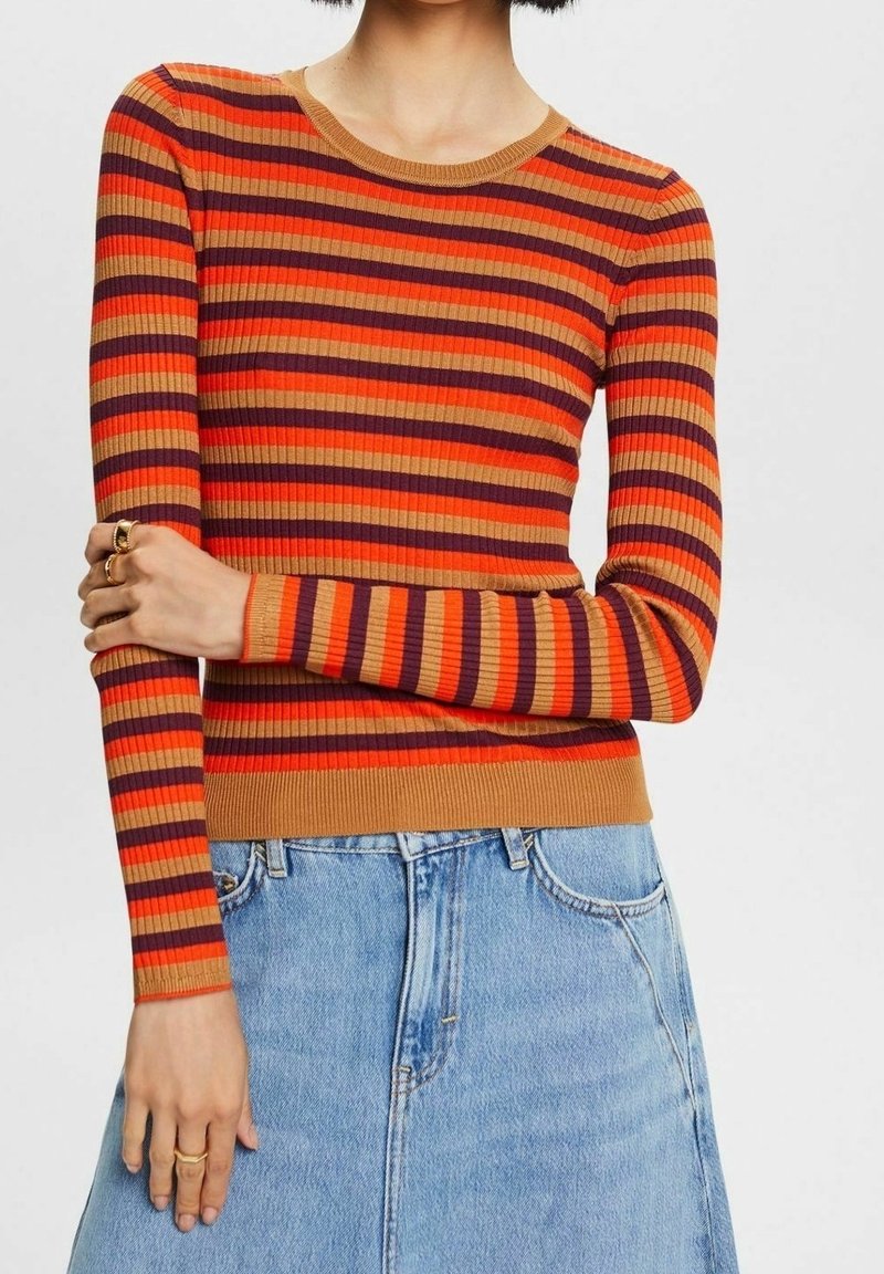 Femme portant un pull côtelé à manches longues avec des rayures horizontales orange, rouges et violettes, rentré dans un jean en denim bleu clair.