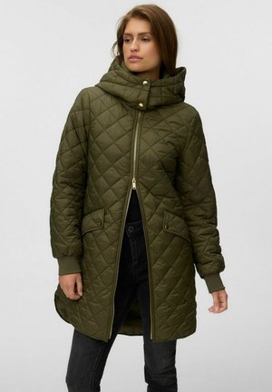 Femme portant une longue veste matelassée vert olive avec col montant, fermeture éclair frontale et poches à rabat, portée avec un jean noir.