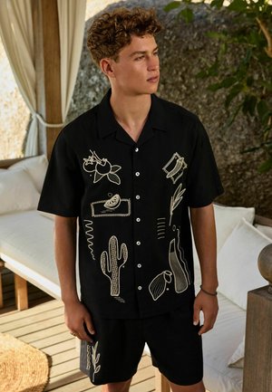 Tânăr cu păr creț, purtând o cămașă neagră cu nasturi și pantaloni scurți cu modele abstracte bej de plante și fructe, stând în interior lângă o canapea.