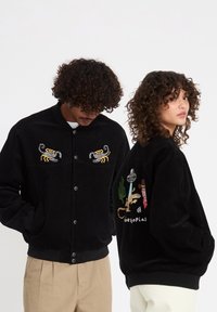 Deux personnes aux cheveux bouclés portent des vestes noires ; la veste avant est brodée d'un scorpion, tandis que la veste arrière présente un motif de cactus et de cow-boy.