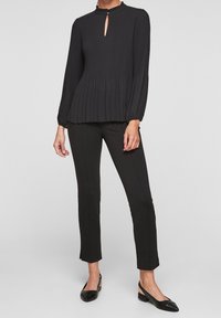 Svart plisserad blus med rund hals och nyckelhål, kombinerad med svarta slim-fit byxor och platta svarta skor.