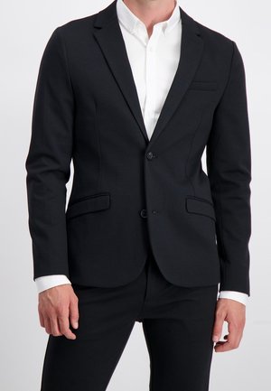 Hombre vistiendo un traje negro ajustado con chaqueta y pantalones, y una camisa blanca de vestir, posando frente a un fondo claro y sencillo.