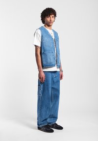 Chaleco de mezclilla azul claro con dos bolsillos frontales, superpuesto a una camiseta blanca. Combinado con pantalones de mezclilla azul holgados y zapatillas negras.