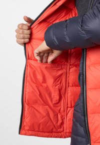 Hände stecken in eine große Innentasche einer orange gefütterten marineblauen Steppjacke, der Reißverschluss ist teilweise geöffnet.