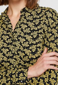 Svart skjorta med ett gult blommönster, knäppdesign, krage och texturerad tyg. Synlig manschettdetaljering.