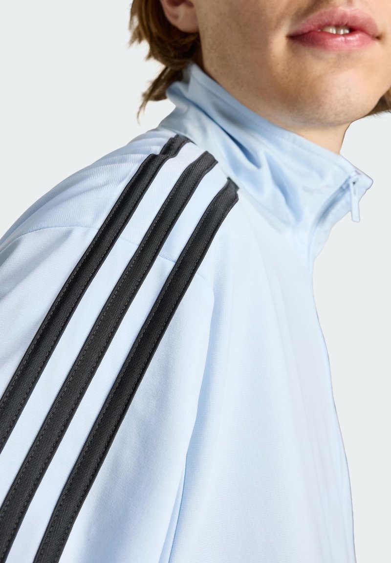 Primo piano di una persona che indossa una giacca azzurro chiaro con tre strisce nere sulla spalla e un colletto con zip.