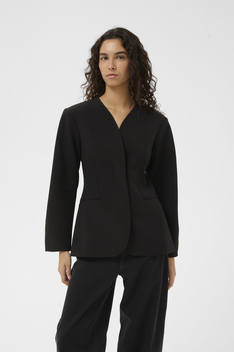 Svart långärmad kavaj med v-ringning, med figursydd stil, två framfickor och slät textur. Kombineras med lösa svarta byxor.