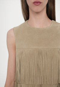 Top fără mâneci din suede, de culoare bej, având o secțiune superioară netedă și detalii cu franjuri verticale în partea inferioară. Design simplu al gâtului.