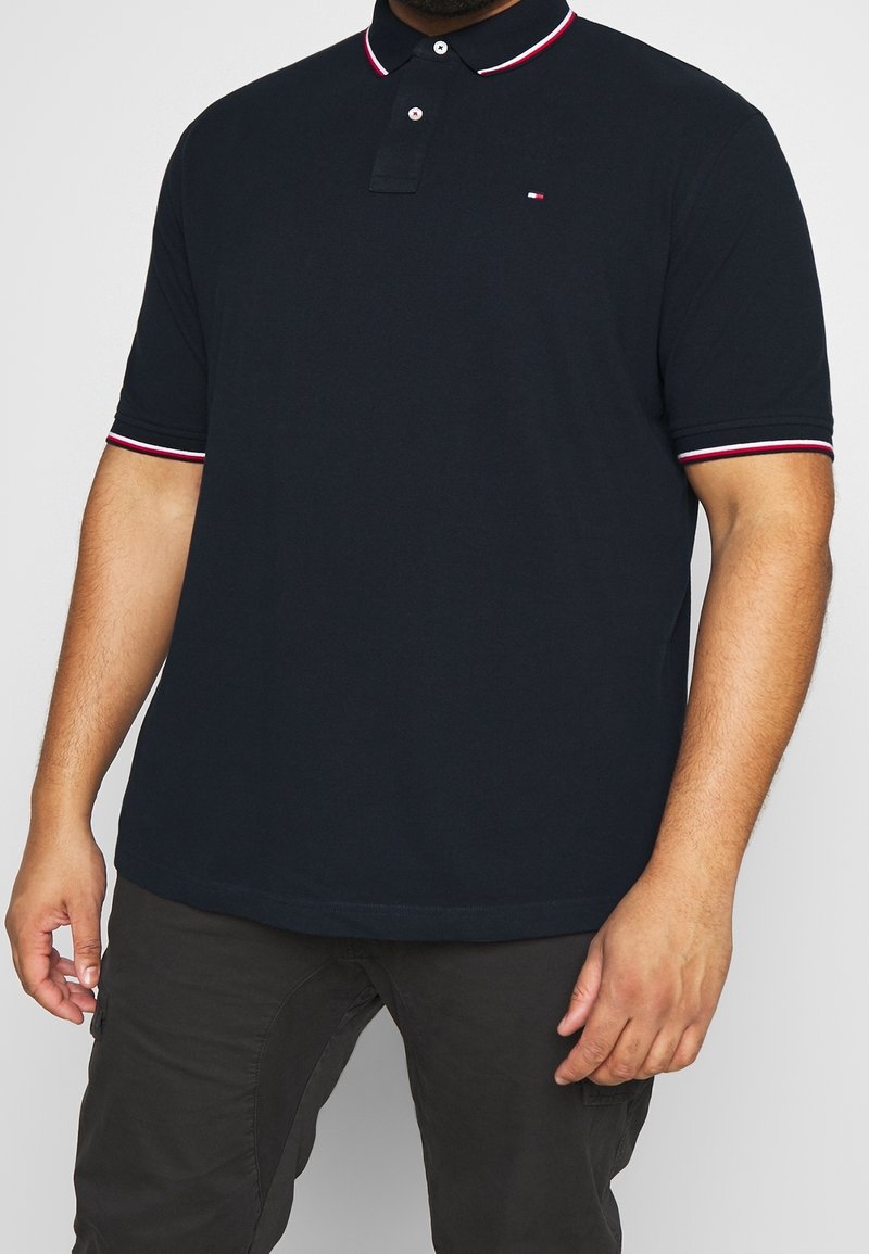 Homme portant un polo bleu marine avec des bordures rouges et blanches sur le col et les manches, associé à un pantalon foncé, debout devant un fond gris.