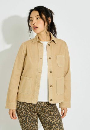 Mujer con una chaqueta abotonada beige sobre una camisa blanca y pantalones con estampado de leopardo, mirando ligeramente de lado contra un fondo blanco.