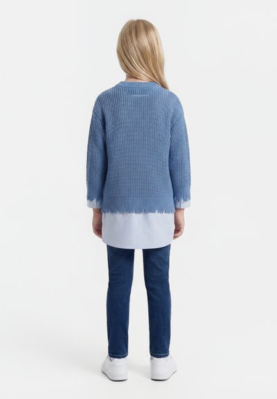 Dieťa s dlhými blond vlasmi stojí chrbtom, má na sebe modrý pletený sveter, svetlomodrú košeľu, tmavé džínsy a biele tenisky.