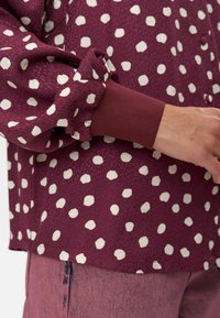 Gros plan sur une main portant un blouse bordeaux à pois blancs avec un poignet côtelé, associée à un jean bordeaux délavé.