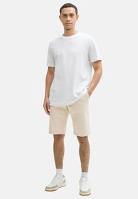 Weißes Baumwoll-T-Shirt, kurze Ärmel, lockere Passform. Hellbeige Shorts, knielang, mit flachem Fuß. Weiße Sneaker mit beigen Akzenten, gerippte Bündchen.
