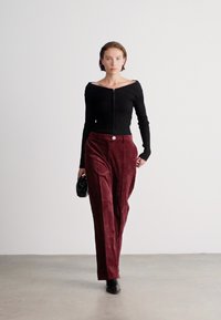 Melna ribota blūze bez pleca ar rāvējslēdzēju, apvienota ar bordo aksesuāru platām biksēm, kurām ir poga. Modele tur melnu somiņu.