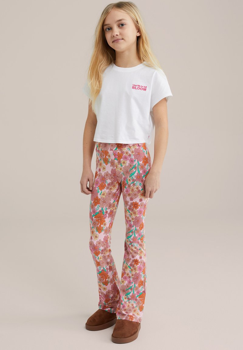 Balts īsais T-krekls ar uzrakstu "Dzīvo dzīvi pilnīgā ZIEDĒšanā". Rozā ziedu raksta zvīņveida bikses ar oranžiem un tirkīza akcentiem. Brūnas čības.