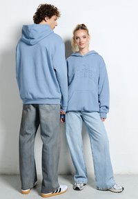 Młody mężczyzna i kobieta w jasnoniebieskich bluzach z kapturem i szerokich dżinsach stojący przy białej ścianie, trzymając się za ręce.